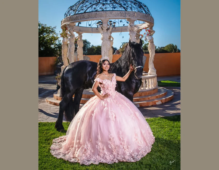 Quinceañera posing with a black horse beside an ornate garden gazebo, wearing a blush pink floral ball gown and crystal tiara