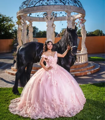 Quinceañera posing with a black horse beside an ornate garden gazebo, wearing a blush pink floral ball gown and crystal tiara