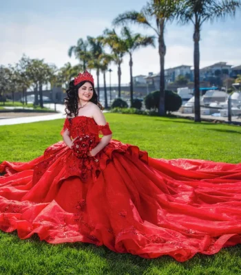 Quinceañera full-length portrait in a red ball gown with a long flowing train, posing on a lawn near a marina