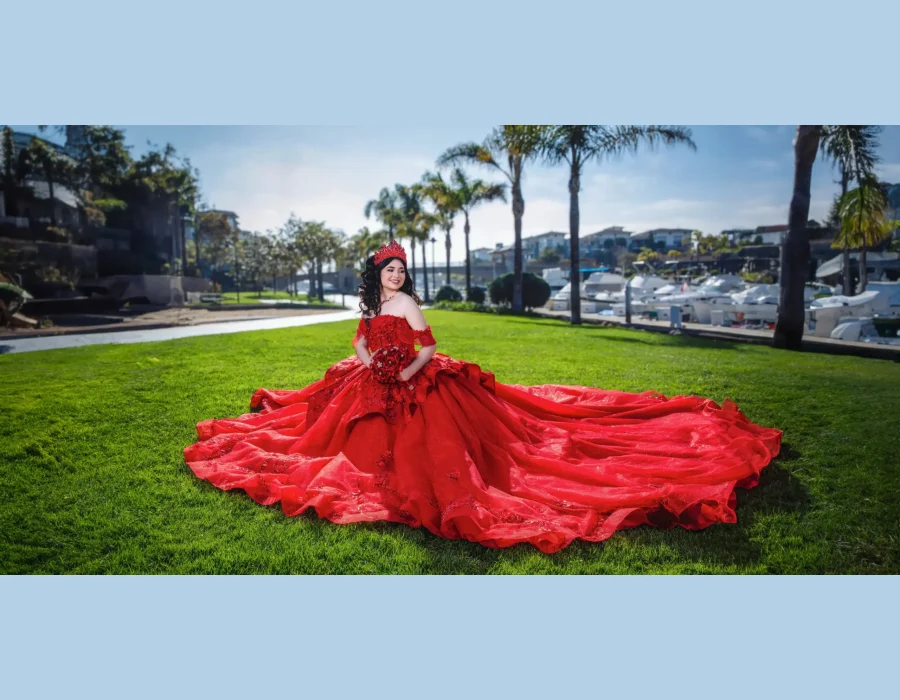 Quinceañera full-length portrait in a red ball gown with a long flowing train, posing on a lawn near a marina