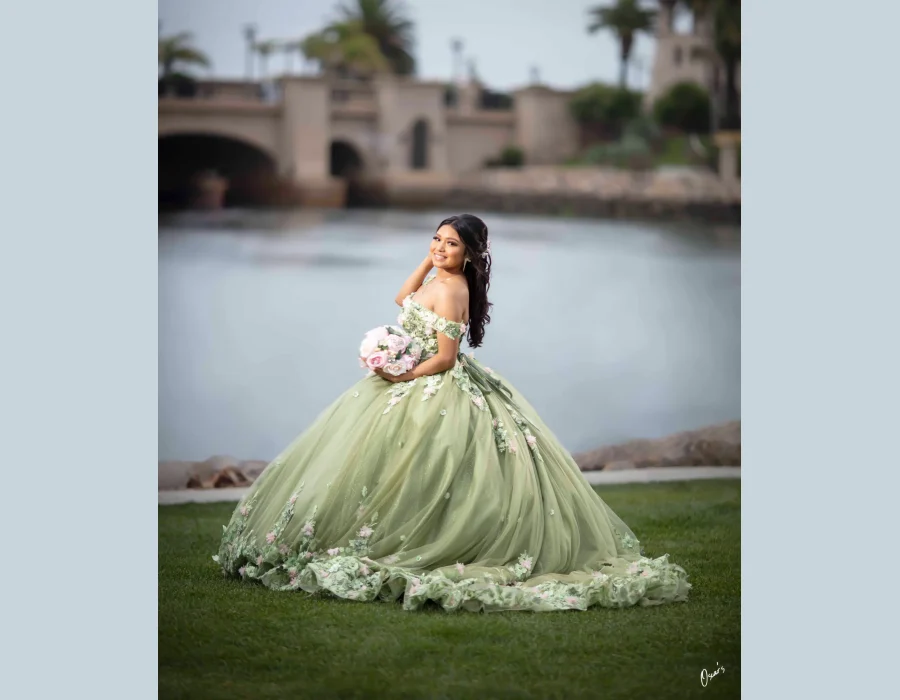 Quinceañera portrait in a sage green floral ball gown with a pink bouquet, posing beside a lake with a bridge in the background