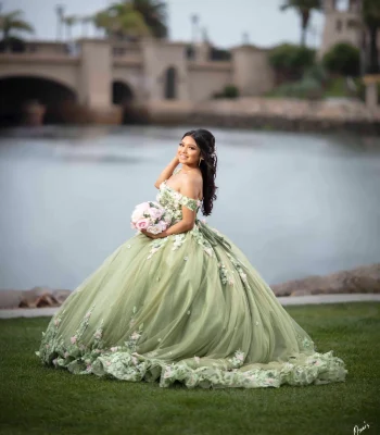 Quinceañera portrait in a sage green floral ball gown with a pink bouquet, posing beside a lake with a bridge in the background