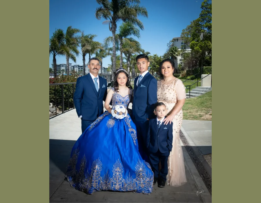 Quinceañera family portrait in a royal blue gown with gold embroidery, surrounded by her parents, brother, and little brother on a palm-lined walkway
