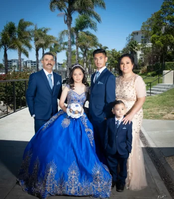 Quinceañera family portrait in a royal blue gown with gold embroidery, surrounded by her parents, brother, and little brother on a palm-lined walkway