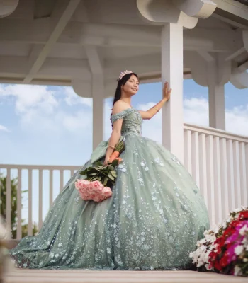Quinceañera portrait in a sage green beaded gown, standing on a white garden gazebo holding a pink rose bouquet