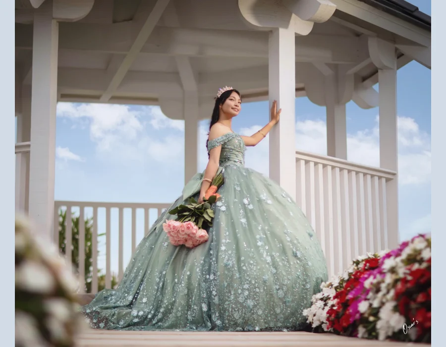 Quinceañera portrait in a sage green beaded gown, standing on a white garden gazebo holding a pink rose bouquet