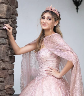 Quinceañera portrait in a blush pink sequined gown with a sheer sparkling cape, posing beside a stone column