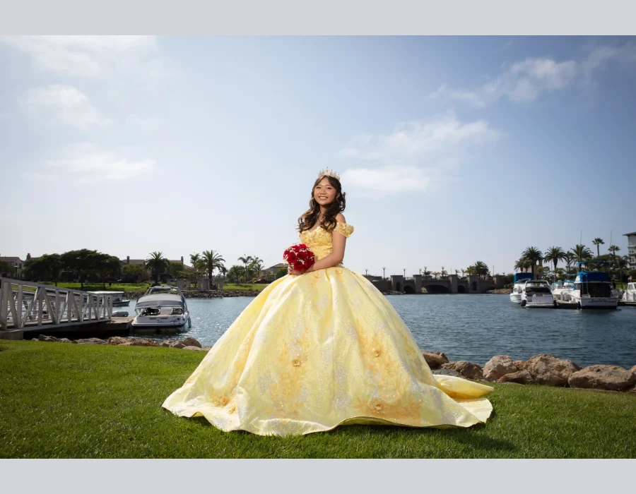 Quinceañera portrait in a golden yellow ball gown holding a red rose bouquet, standing beside a marina with boats and palm trees