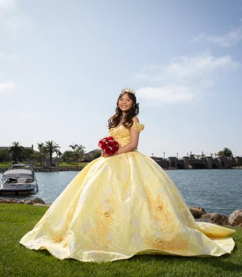 Quinceañera portrait in a golden yellow ball gown holding a red rose bouquet, standing beside a marina with boats and palm trees