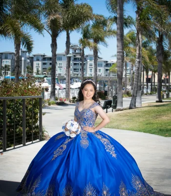 Quinceañera full-length portrait in a royal blue satin gown with gold embroidery, posing on a palm-lined path near the marina