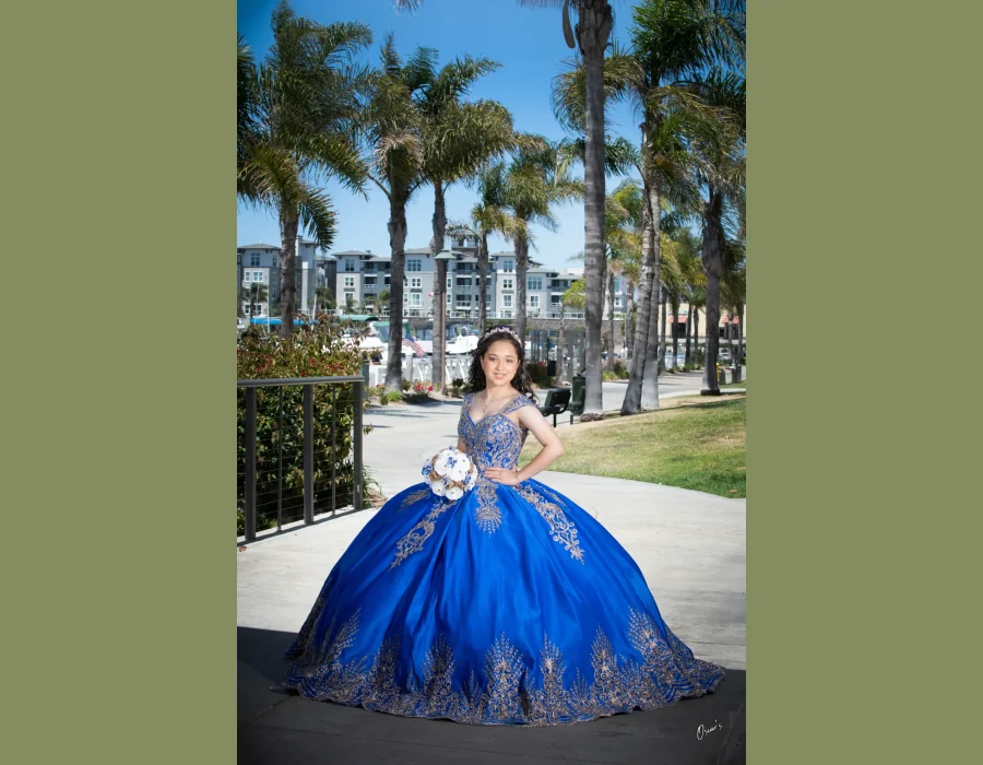 Quinceañera full-length portrait in a royal blue satin gown with gold embroidery, posing on a palm-lined path near the marina