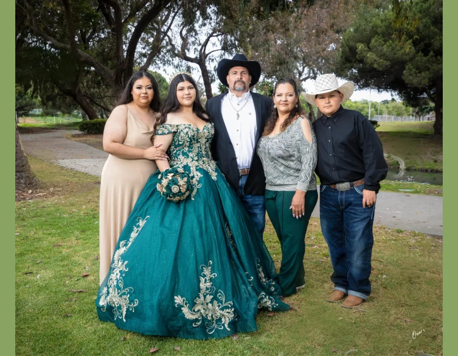 Quinceañera family portrait in an emerald green gown with gold lace, surrounded by parents and siblings in a park setting