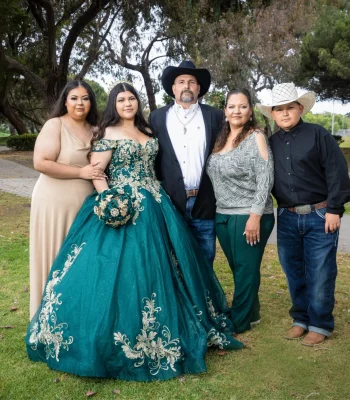 Quinceañera family portrait in an emerald green gown with gold lace, surrounded by parents and siblings in a park setting
