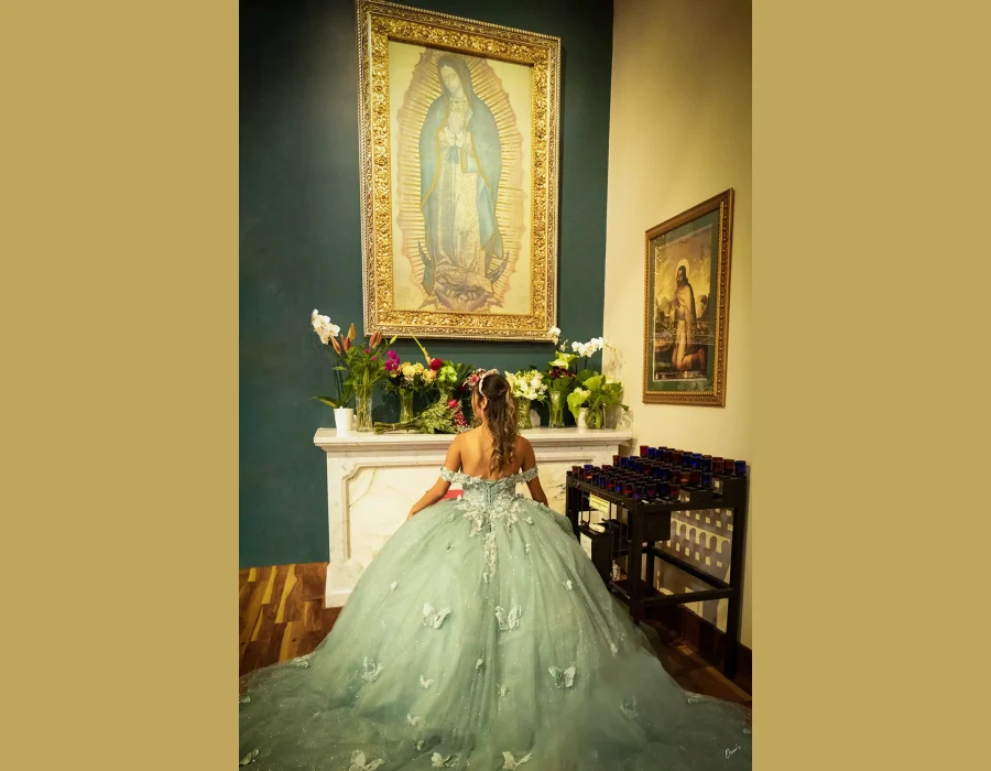 Quinceañera portrait from behind, kneeling before a devotional altar with framed religious art in a sage green butterfly-embellished gown