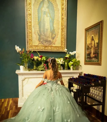 Quinceañera portrait from behind, kneeling before a devotional altar with framed religious art in a sage green butterfly-embellished gown