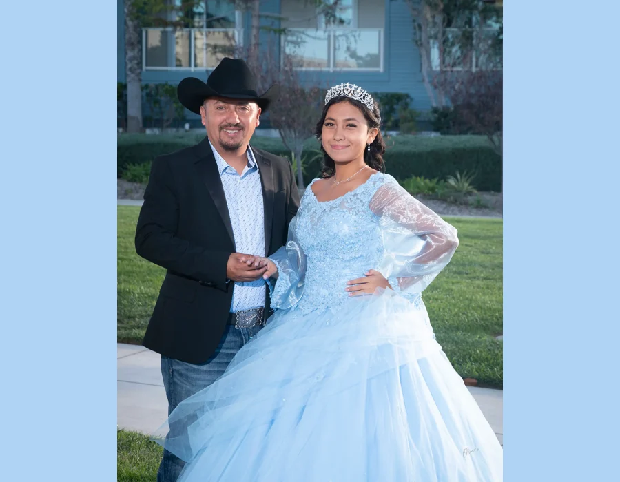Quinceañera portrait with her father in a light blue lace gown with sheer sleeves and a silver tiara, standing on a lawn in front of a house