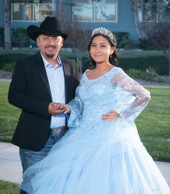 Quinceañera portrait with her father in a light blue lace gown with sheer sleeves and a silver tiara, standing on a lawn in front of a house