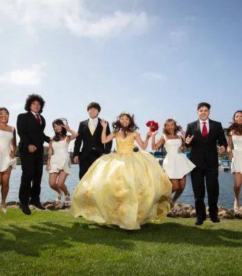 Quinceañera group portrait in a golden yellow ball gown, jumping for joy with friends in black suits and white dresses by the marina