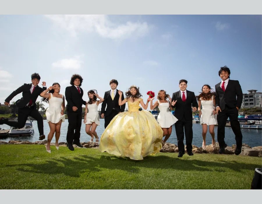 Quinceañera group portrait in a golden yellow ball gown, jumping for joy with friends in black suits and white dresses by the marina