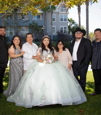Quinceañera family portrait in a pastel floral gown, surrounded by parents and relatives under a tree near an apartment building
