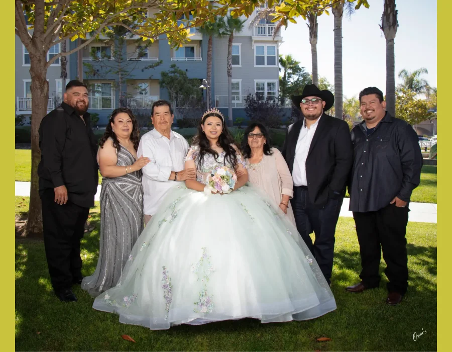 Quinceañera family portrait in a pastel floral gown, surrounded by parents and relatives under a tree near an apartment building