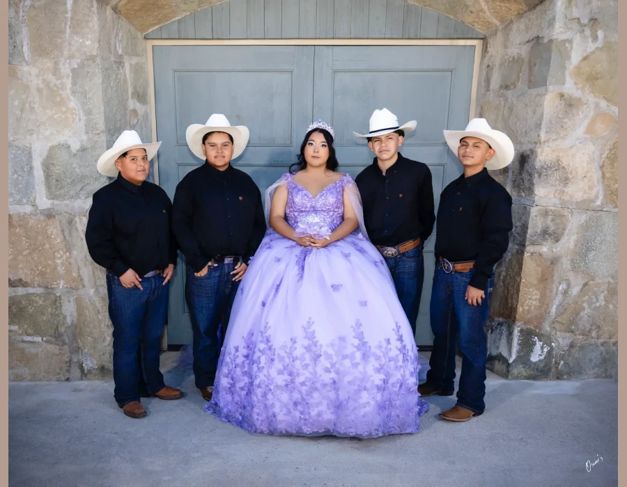 Quinceañera portrait in a lavender floral gown, standing with four young men in cowboy hats in front of stone archway doors