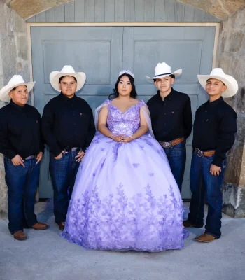 Quinceañera portrait in a lavender floral gown, standing with four young men in cowboy hats in front of stone archway doors