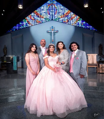 Quinceañera family portrait in a blush pink floral gown, standing before a stained glass window and cross inside a church