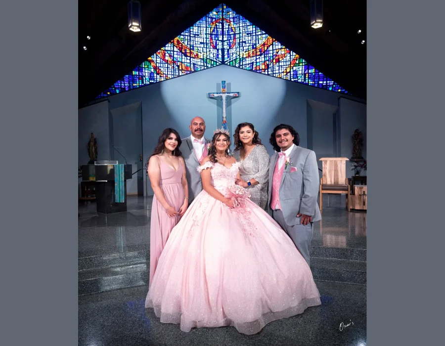 Quinceañera family portrait in a blush pink floral gown, standing before a stained glass window and cross inside a church
