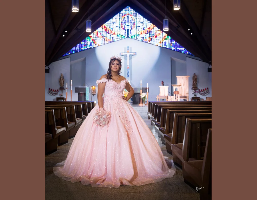 Quinceañera portrait in a blush pink floral gown, standing in the aisle of a church with a stained glass window overhead