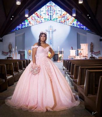 Quinceañera portrait in a blush pink floral gown, standing in the aisle of a church with a stained glass window overhead