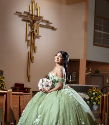 Quinceañera portrait in a sage green floral gown, smiling beside a gold crucifix inside a church