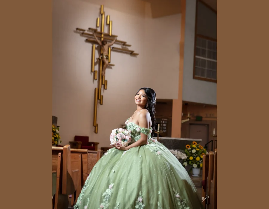 Quinceañera portrait in a sage green floral gown, smiling beside a gold crucifix inside a church
