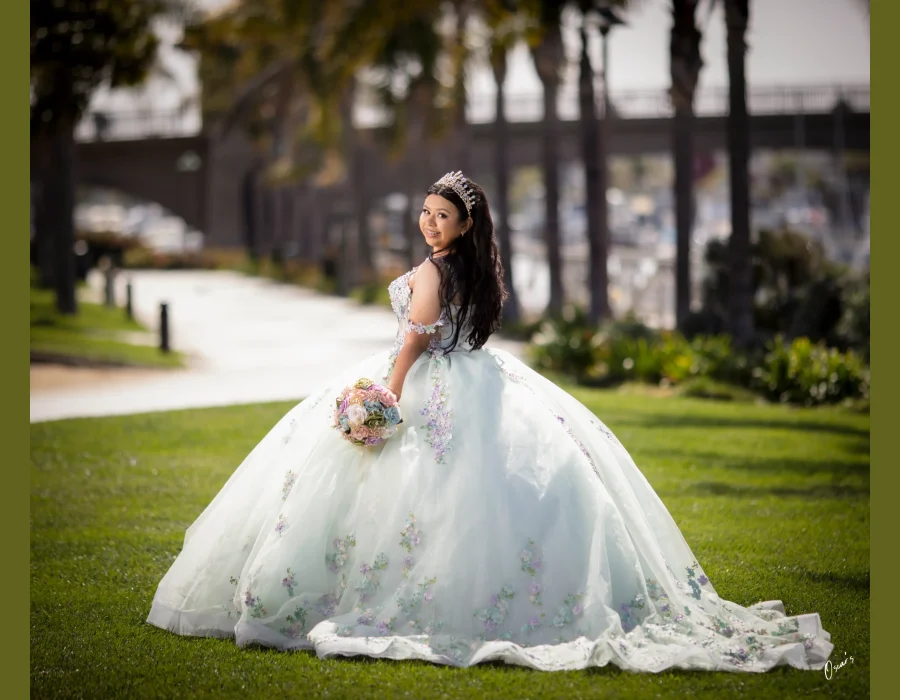 Quinceañera portrait in a pastel floral gown with a crystal tiara, looking back over her shoulder on a path near a marina