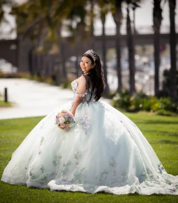 Quinceañera portrait in a pastel floral gown with a crystal tiara, looking back over her shoulder on a path near a marina