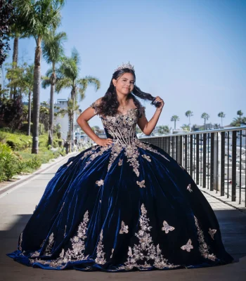 Quinceañera portrait in a navy blue velvet gown with gold floral appliqués, playing with her hair on a waterfront promenade