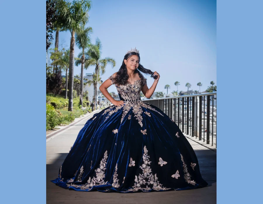 Quinceañera portrait in a navy blue velvet gown with gold floral appliqués, playing with her hair on a waterfront promenade