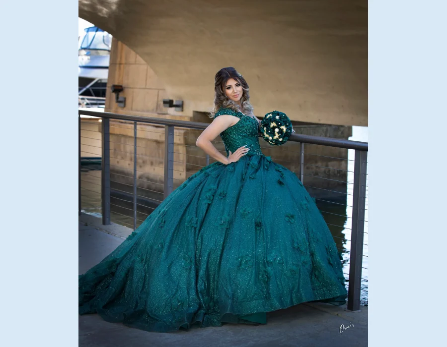 Quinceañera portrait in an emerald green gown, posing on a bridge railing near a marina