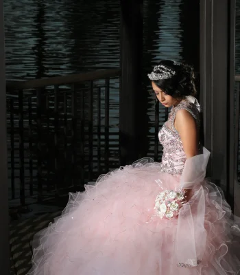 Quinceañera nighttime portrait in a blush pink ruffled gown with a crystal tiara, standing by a dark waterfront railing