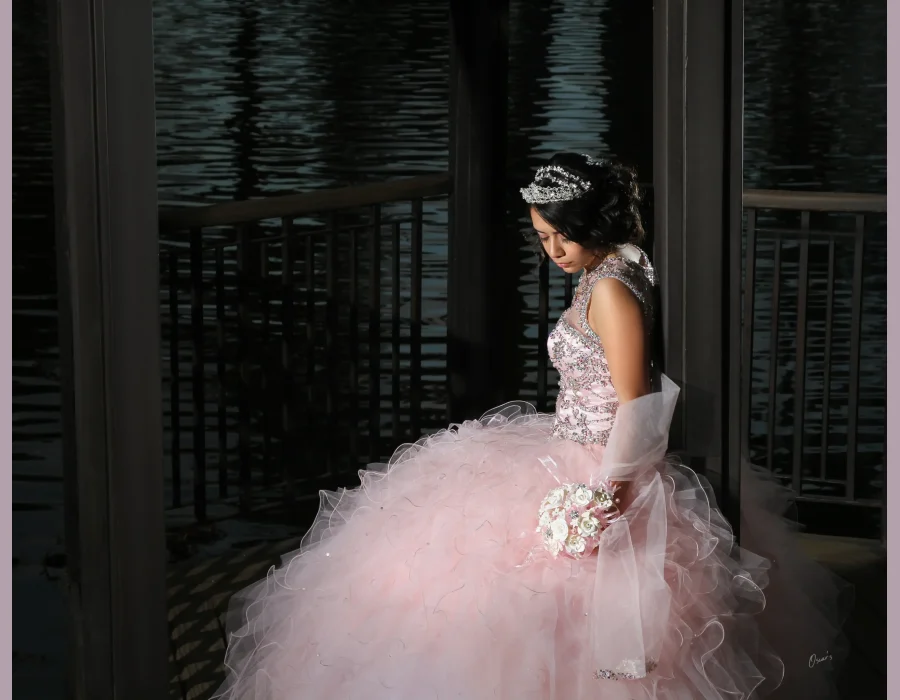 Quinceañera nighttime portrait in a blush pink ruffled gown with a crystal tiara, standing by a dark waterfront railing