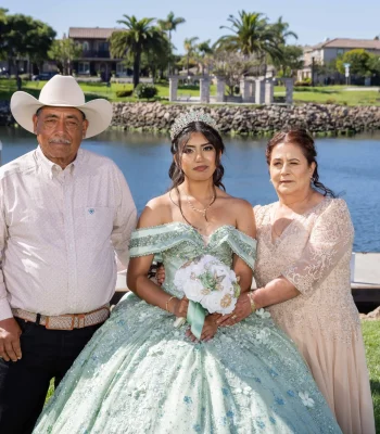 Quinceañera family portrait in a sage green beaded gown, standing with her parents beside a lake