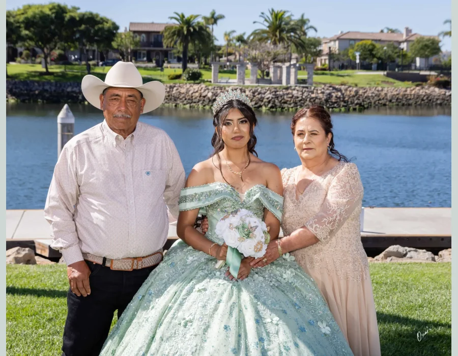 Quinceañera family portrait in a sage green beaded gown, standing with her parents beside a lake