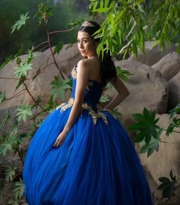 Quinceañera portrait in a royal blue gown with gold floral embroidery, posing with hands on hips against greenery