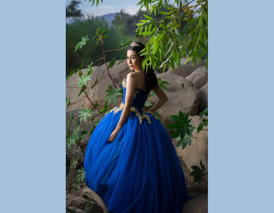 Quinceañera portrait in a royal blue gown with gold floral embroidery, posing with hands on hips against greenery