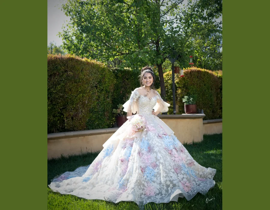 Quinceañera portrait in an ivory beaded gown with sheer bell sleeves, smiling in a garden with hummingbird feeders