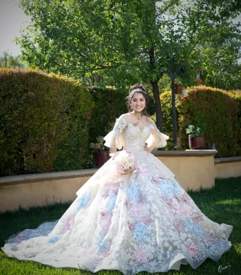 Quinceañera portrait in an ivory beaded gown with sheer bell sleeves, smiling in a garden with hummingbird feeders