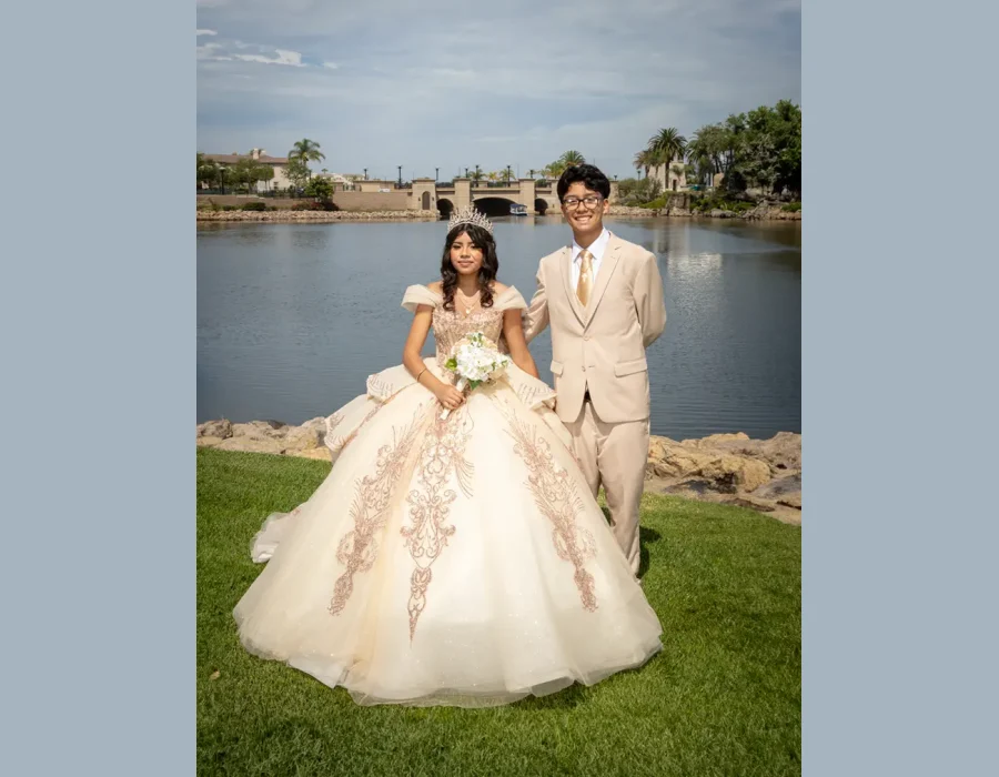 Quinceañera portrait with her escort in a champagne gown with gold embroidery, standing by a lake in matching tan suit