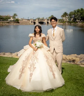 Quinceañera portrait with her escort in a champagne gown with gold embroidery, standing by a lake in matching tan suit