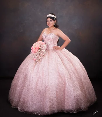 Quinceañera studio portrait in a blush pink glitter gown, smiling and holding a pink rose bouquet against a dark backdrop