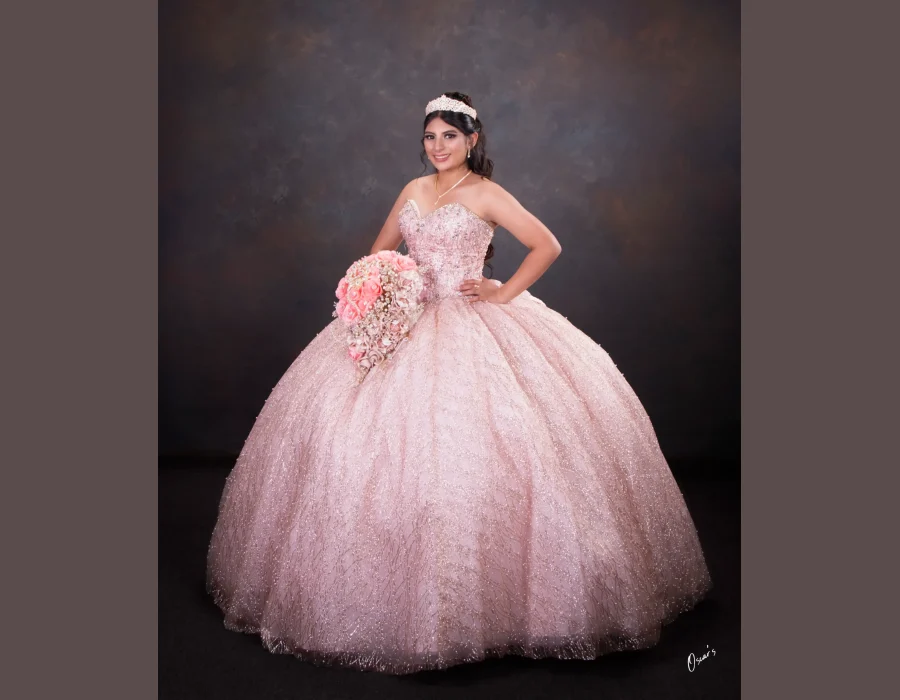 Quinceañera studio portrait in a blush pink glitter gown, smiling and holding a pink rose bouquet against a dark backdrop